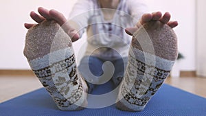 A woman in warm knitted socks does morning exercises, while sitting stretches her fingers up to her toes.