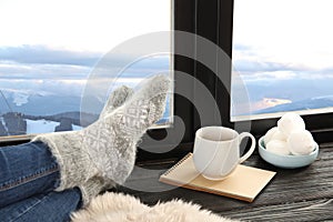 Woman in warm socks with hot drink enjoying view of winter mountain landscape from window
