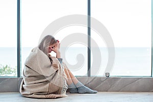 Woman in warm socks sitting on a floor near large window wrapped in a blanket, holding her head, having a strong headache