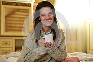 Woman in warm sweater with cup of hot drink on bed at home