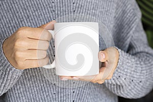 Woman in warm sweater holding white mug in hands. Winter cup Mockup for Christmas individual gifts design