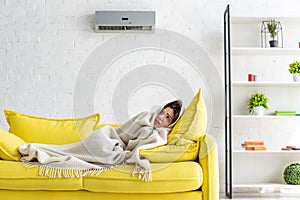 Woman warming with blanket while lying on yellow sofa under air conditioner at home