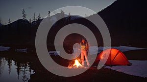 Woman warming hands by the fire in the camping in mountains in the evening