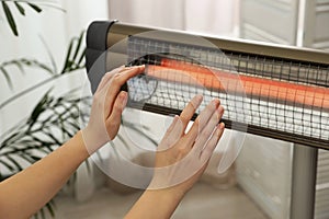 Woman warming hands near electric infrared heater indoors, closeup