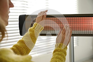 Woman warming hands near electric infrared heater indoors, closeup