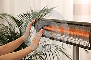 Woman warming hands near electric infrared heater indoors, closeup