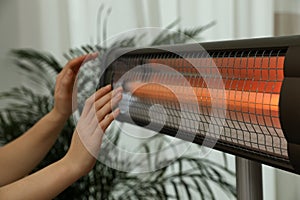 Woman warming hands near electric infrared heater indoors, closeup