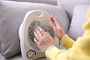 Woman warming hands near fan heater indoors, closeup