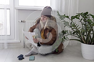 A woman warmly dressed in a brown jacket and hat is sitting near a heating appliance with bills in her hands, money and