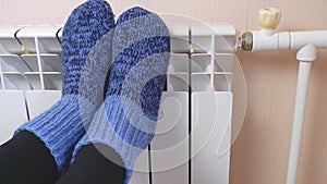 A woman warms her feet by the radiator on a cold winter day in blue woolen socks. Central heating system. Expensive