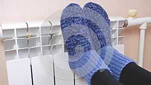 A woman warms her feet by the radiator on a cold winter day in blue woolen socks. Central heating system. Expensive