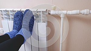 A woman warms her feet by the radiator on a cold winter day in blue woolen socks. Central heating system. Expensive