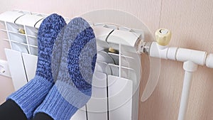 A woman warms her feet by the radiator on a cold winter day in blue woolen socks. Central heating system. Expensive