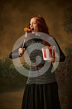 Woman warrior in chainmail and dress eating modern junk food from serving fork against vintage studio background.