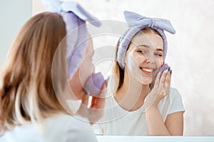 Woman washes her face with natural sponge and soap scrub cosmetic foam in bathroom portrait reflection in mirror