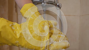 A woman washes her hands in yellow rubber gloves in the sink under the tap with water. Housewife washes soap with