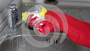 Woman washes a metal faucet with sponge in the kitchen in red gloves. Hands close-up.