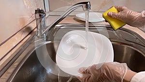 Woman washes white plate in kitchen sink. Close up of faucet and female hand in gloves. Water streaming. Brown