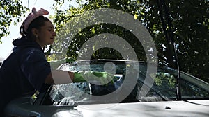 A woman washes the windshield of a car with a soapy sponge in the yard of her house in summer