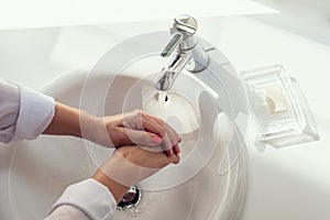 Woman washing hands rubbing with soap in white sink in bathroom with sunlight from the window. Protection from bacteria and virus.