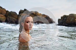 Woman with waterproof makeup submerges in ocean, stones backdrop. Female tours seascape, enduring swimwear stylish