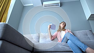 Woman waving paper sitting on couch, suffering heat, faulty air conditioning