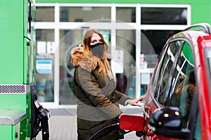 Woman wear medical mask at self-service gas station, hold fuel nozzle, refuel the car with petrol during corona virus