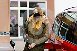 Woman wear medical mask at self-service gas station, hold fuel nozzle, refuel the car with petrol during corona virus
