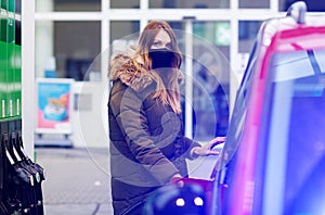 Woman wear medical mask at self-service gas station, hold fuel nozzle, refuel the car with petrol during corona virus