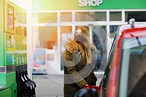 Woman wear medical mask at self-service gas station, hold fuel nozzle, refuel the car with petrol during corona virus