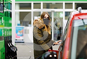 Woman wear medical mask at self-service gas station, hold fuel nozzle, refuel the car with petrol during corona virus