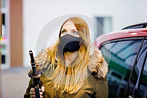 Woman wear medical mask at self-service gas station, hold fuel nozzle, refuel the car with petrol during corona virus