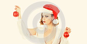 Woman wear sexy dress and santa hat hold decorative balls on white background. Girl with makeup and hairstyle ready for