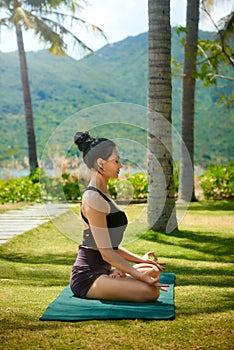 Woman wearing active wear doing yoga practice meditating in park nature, lotus position. Meditation in sunny day, inner balance