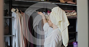 Woman wearing a bathrobe and a towel on her head choosing clothes from rack with hangers inside room. Lady looks to