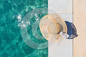 Woman wearing big summer sun hat relaxing on pier by clear turquoise sea.