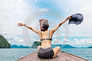 Woman wearing bikini sitting on the boat with hands up and holding beachhat.