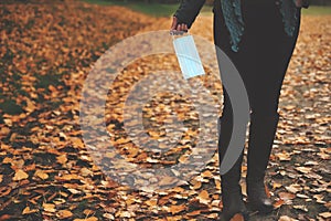 woman wearing black jeans and boots holding a light blue disposable protective face mask stands in autumn colored surrounding