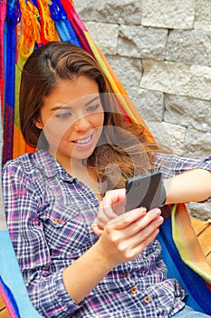 Woman wearing casual clothes holding mobile phone lying in hammock smiling, raising arm looking at smart watch, grey