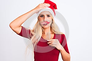 Woman wearing Christmas Santa hat holding fanny mustache over isolated white background stressed with hand on head, shocked with