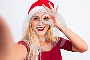Woman wearing Christmas Santa hat make selfie by camera over isolated white background with happy face smiling doing ok sign with