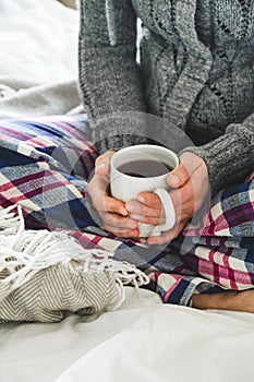 Woman wearing cozy pyjamas and gray cardigan drinking tea on a bed
