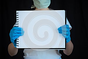 Woman wearing face mask and surgical gloves, holding blank note pad with black background