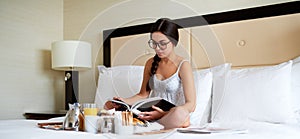 Woman wearing glasses and nightgown reading book in bed.