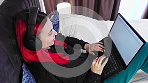 Woman wearing headphones with microphone sits on a bed and works remotely from her cozy bedroom