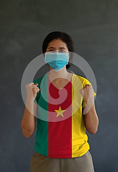 Woman wearing hygienic mask and wearing Cameroon flag colored shirt and standing with raised both fist on dark wall background