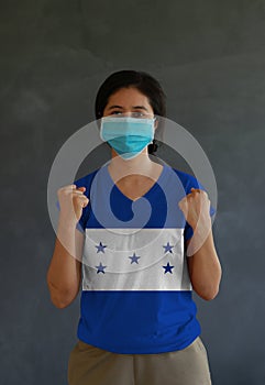 Woman wearing hygienic mask and wearing Honduras flag colored shirt and standing with raised both fist on dark wall background