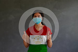 Woman wearing hygienic mask and wearing Tajiks flag colored shirt with thumbs up with both hands on dark wall background