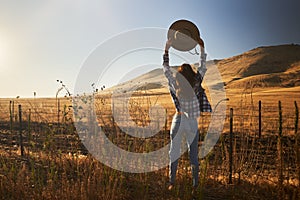 Woman wearing jeans and flannel from behind looking at view of rural california landscape holding up hat in air