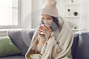 Woman wearing a knitted sweater and hat sitting on the sofa wrapped in a plaid drinking hot coffee.